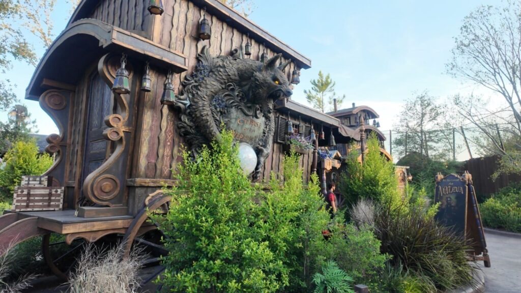 A rustic wooden wagon decorated with lanterns and carvings features a large wolf’s head sculpture with glowing yellow eyes and a crystal ball at its base. Surrounded by greenery, the structure has an eerie, carnival-like vibe, with a chalkboard-style sign reading “Madame” set up nearby, hinting at a fortune-telling or haunted attraction.