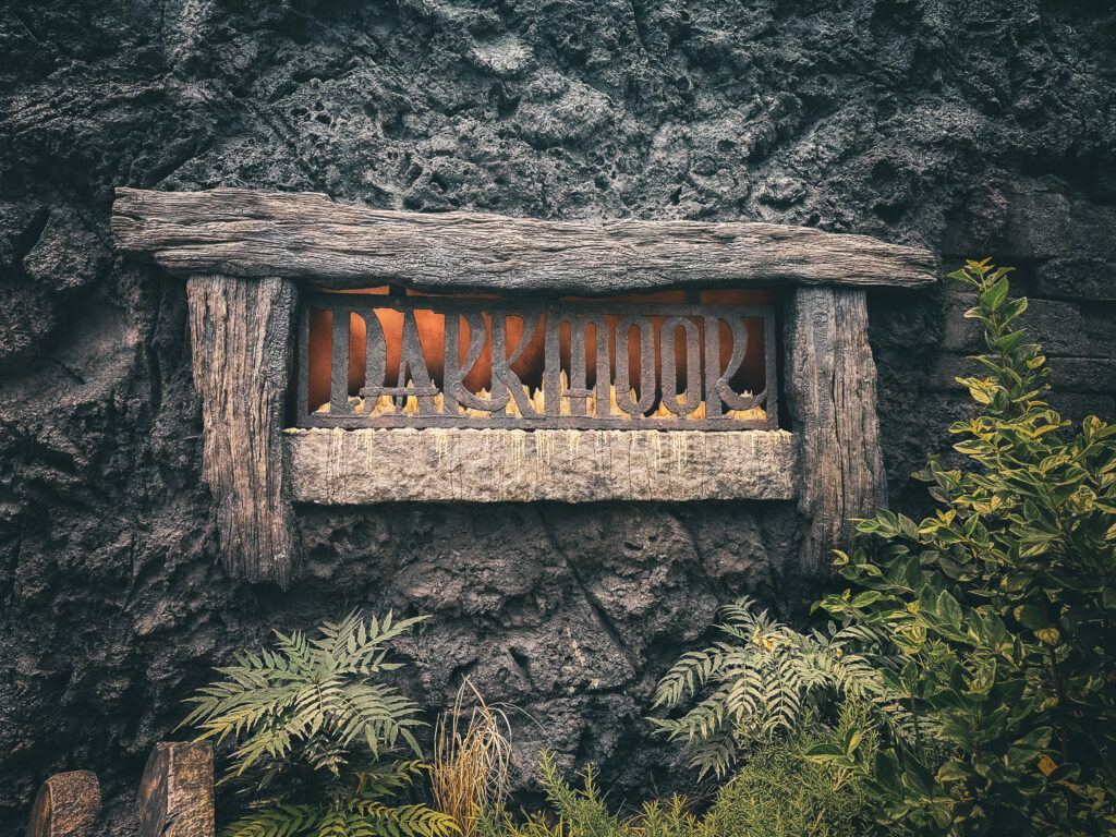 A weathered wooden sign framed in rough beams reads “DARKMOOR,” mounted against a textured stone wall. Green ferns and leafy plants grow at the base, adding contrast to the dark, rugged background.