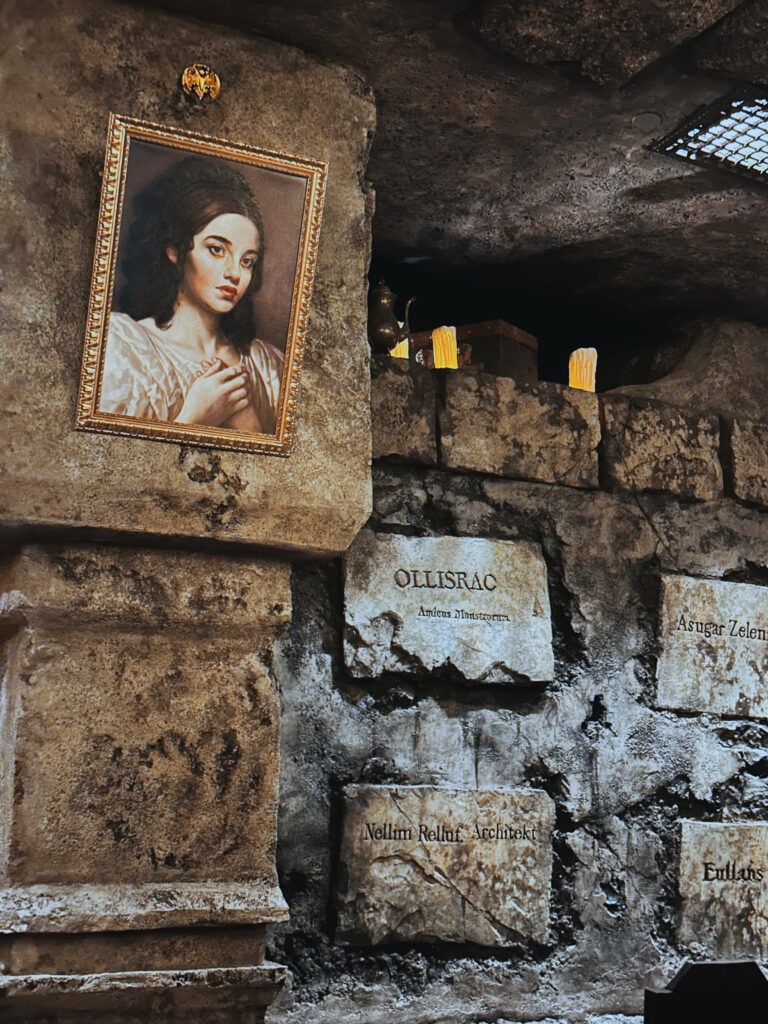 A dim stone wall features weathered plaques with Latin-like inscriptions and a framed portrait of a woman with dark hair in a white gown. Above the plaques, flickering candles and a brass teapot sit on a ledge, adding to the mysterious, old-world atmosphere. The setting feels like a crypt or hidden chamber.