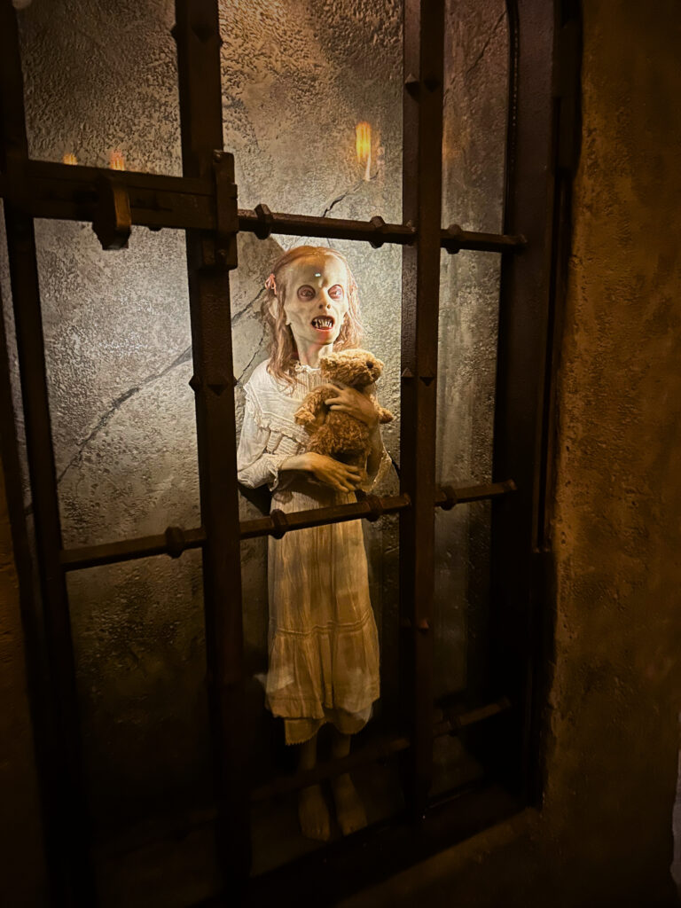 A creepy animatronic figure of a pale, undead-looking girl with glowing eyes stands behind iron bars, clutching a worn teddy bear. She wears a tattered white nightgown and is lit dramatically against a cracked stone wall, creating a chilling, haunted asylum vibe.