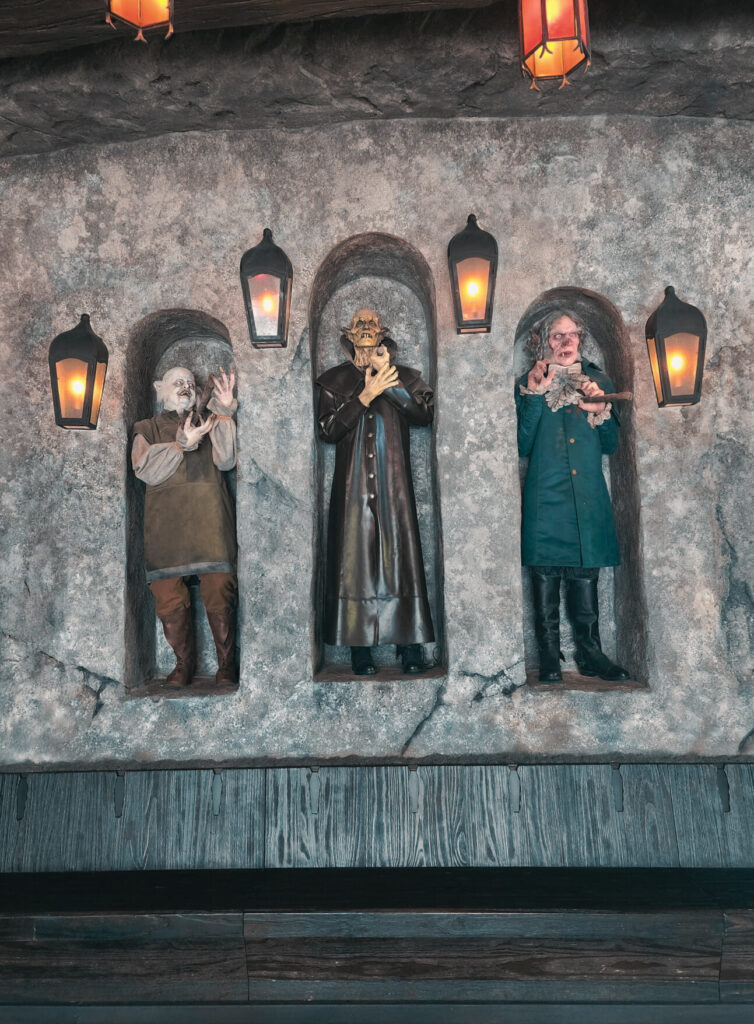 Three eerie statues are set into stone alcoves, each lit by glowing lanterns. The figures include ghoulish characters with pale faces and unsettling expressions, dressed in period-style clothing. The dim lighting and rough stone wall enhance the spooky, haunted atmosphere.
