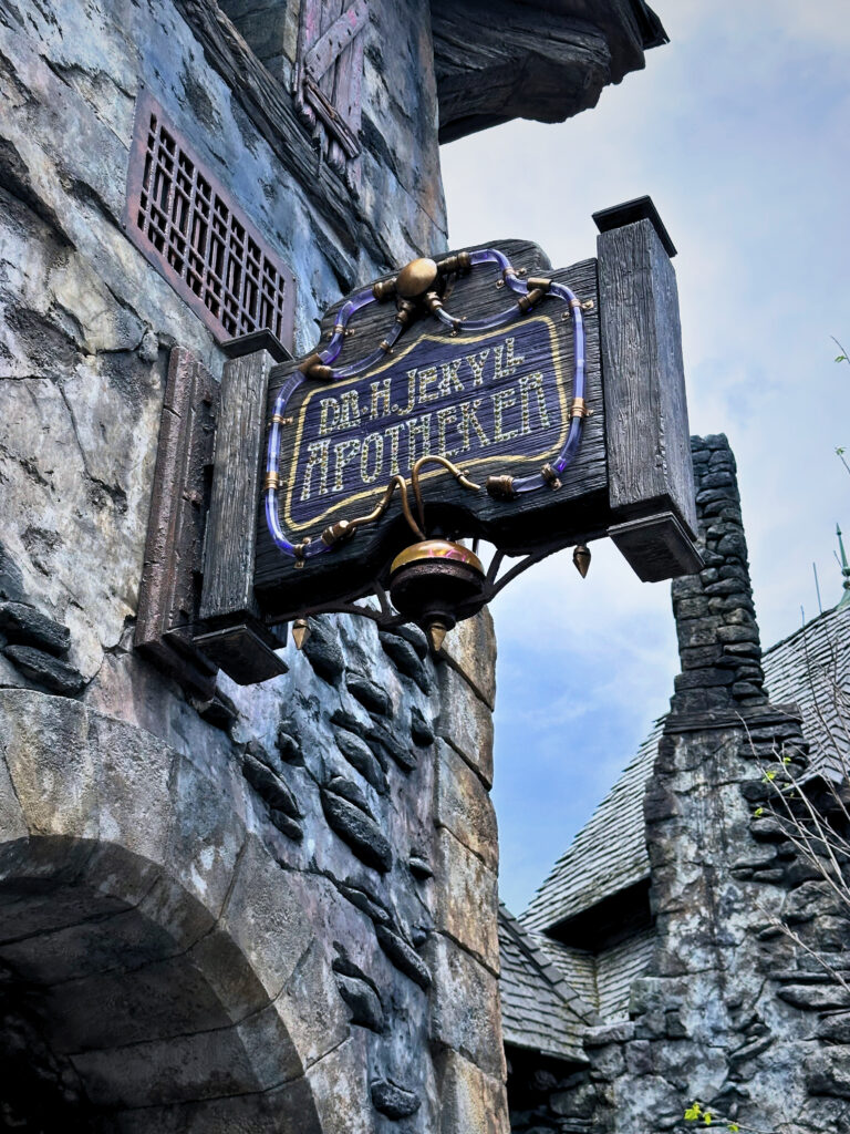 A weathered wooden sign reading "Dr. H. Jekyll Apotheker" hangs from a medieval-style stone building, surrounded by exposed beams and aged stonework. The sign features ornate details with purple and gold accents, adding a gothic, fantasy vibe to the scene.
