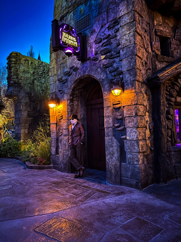 A man in a fedora and suit leans casually against the stone doorway of a dimly lit building marked with a glowing purple sign that reads "Dr. Jekyll's Apothecary." Warm lantern light illuminates the entrance, contrasting with the cool blue evening sky and purple glow from inside the building.
