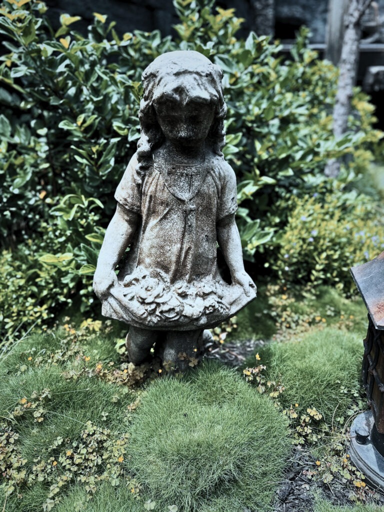 A weathered stone statue of a young girl stands in a garden, holding the hem of her dress filled with carved flowers. The statue is surrounded by lush green plants and mossy ground cover, giving it an aged, slightly mysterious atmosphere.