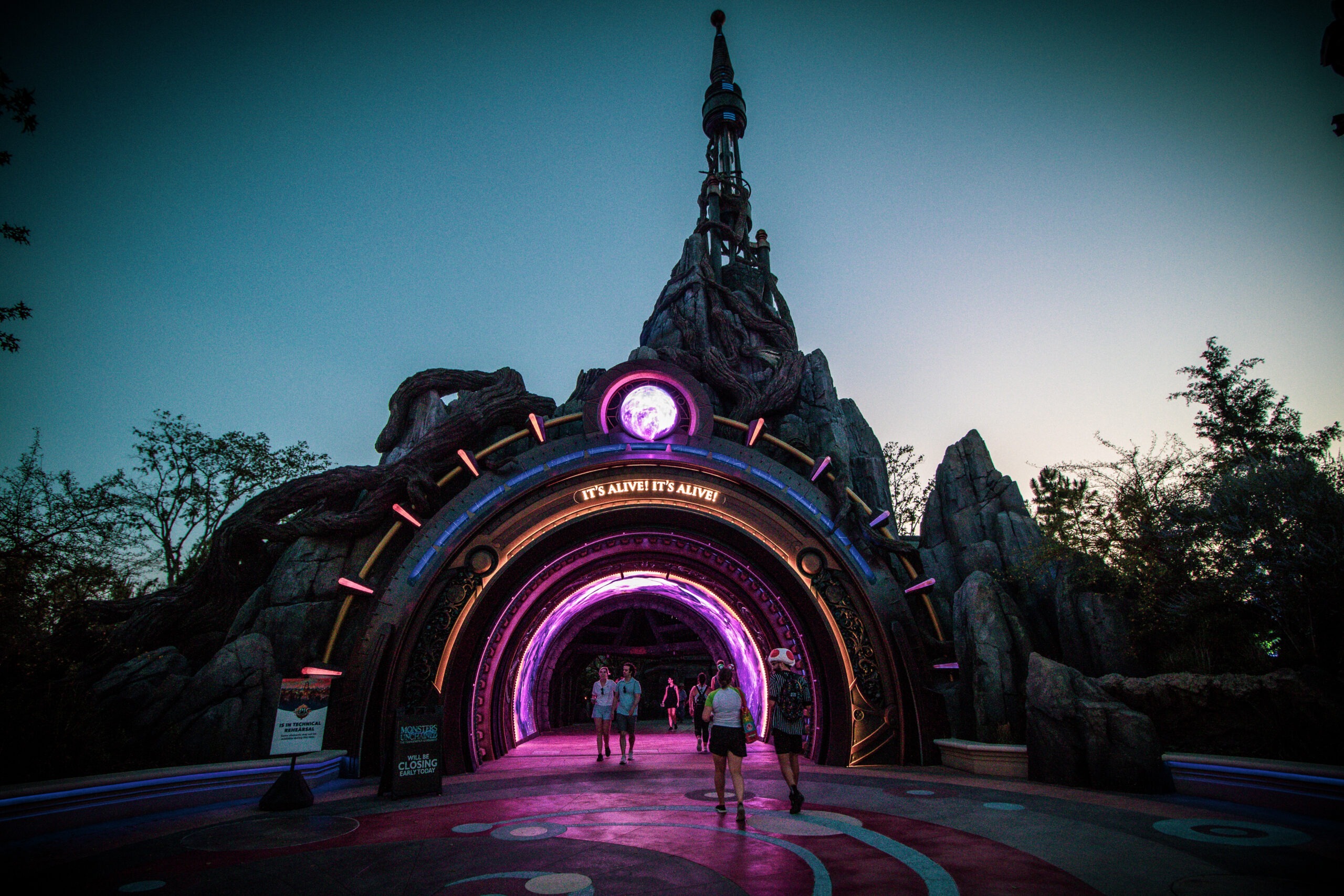 An illuminated archway styled like a futuristic portal glows with purple and pink lights, with the words “IT’S ALIVE! IT’S ALIVE!” displayed above the entrance. Visitors walk through the tunnel, framed by rocky structures and twisted tree roots, giving the scene a mix of sci-fi and fantasy atmosphere.