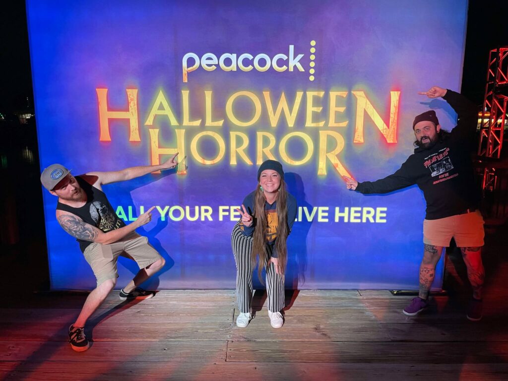 The Fear and Beer podcast hosts pose in front of Peacock's sign for Halloween Horror Nights. 