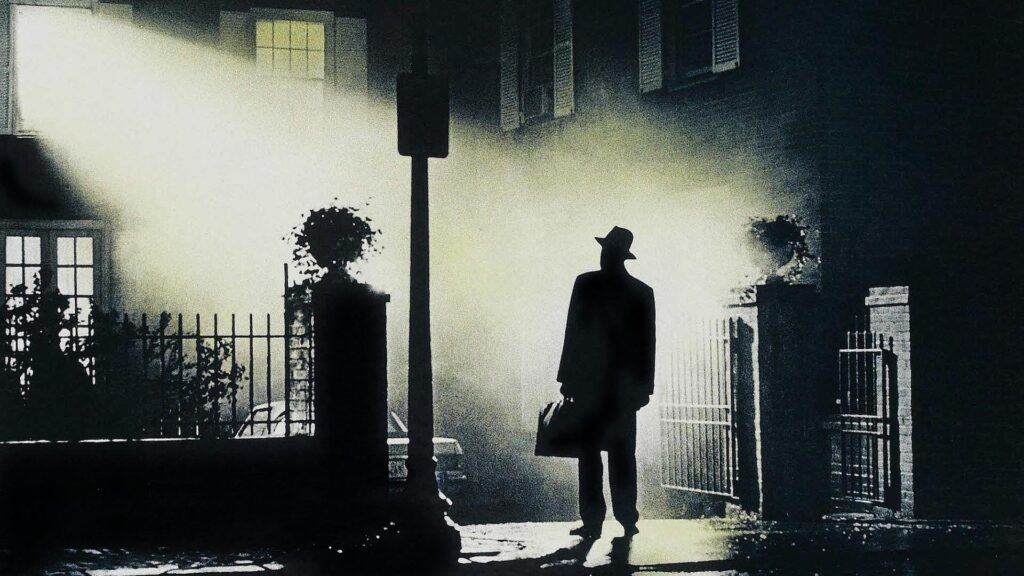 A silhouetted man stands in the light from a window looking at a building. This scene is from the original Exorcist movie.