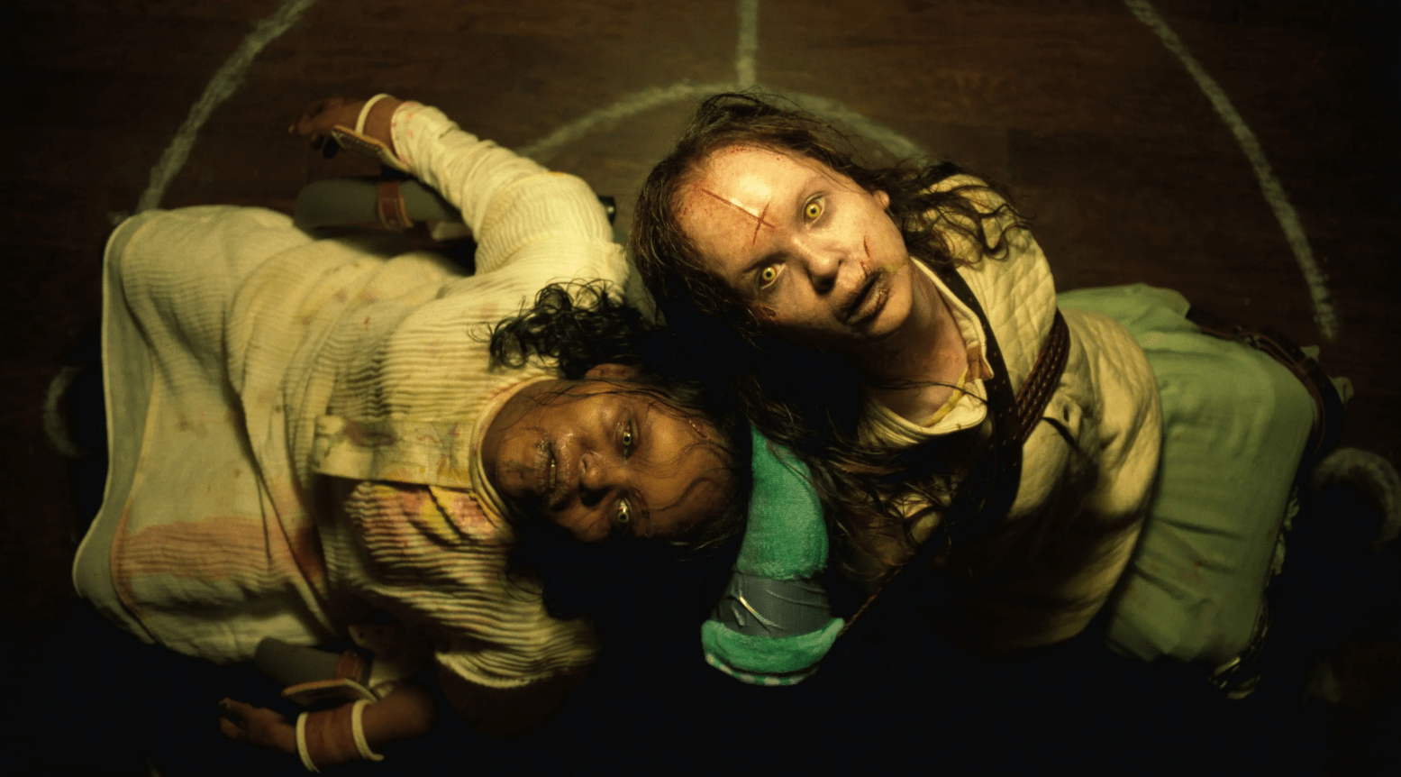 Two young girls with demonic yellow eyes and bloodied faces sit back-to-back, restrained in chairs, inside a ritual circle drawn on a wooden floor. Their heads lean against each other as they look upward with eerie expressions, lit by a sickly yellow light that enhances the horror atmosphere.