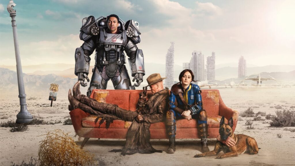 In a barren desert wasteland, three main characters from the Fallout TV series pose around a worn-out orange couch: a woman in a blue Vault-Tec suit sits center, flanked by a trench coat–wearing ghoul with a cowboy hat lounging with his feet across the couch and a loyal German Shepherd resting at her feet. Behind them, a man in bulky power armor stands tall under a pale sky, with crumbling skyscrapers and a retro-futuristic dome in the distance, plus a Route 66 sign and tumbleweed nearby.
