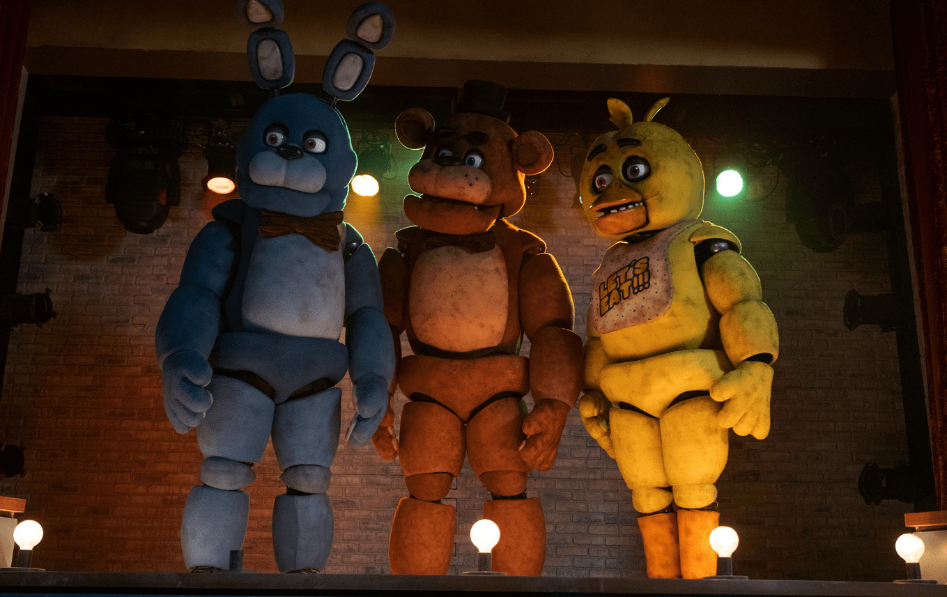 Bonnie, Freddy, and Chica stand side by side on a dimly lit stage, illuminated by warm spotlights in a scene from the *Five Nights at Freddy’s* movie. Each animatronic has a worn, bulky appearance, with Bonnie in blue, Freddy in brown with his signature top hat, and Chica in yellow wearing a bib that reads “LET’S EAT!!!”. The brick wall and stage lighting give the setting a nostalgic yet eerie vibe.