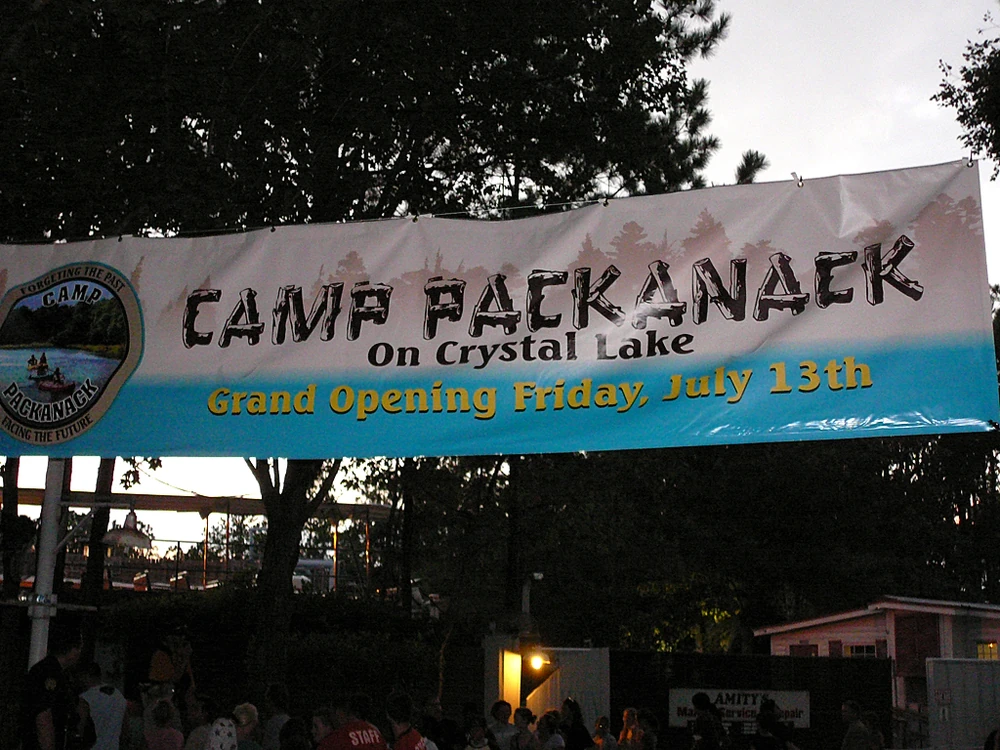 A large outdoor banner reads “Camp Packanack on Crystal Lake – Grand Opening Friday, July 13th” with a camp logo showing canoes on a lake and the tagline “Forgetting the Past, Embracing the Future.” The banner hangs between trees at dusk above a crowd, hinting at a spooky or ironic tone given the Friday the 13th reference.