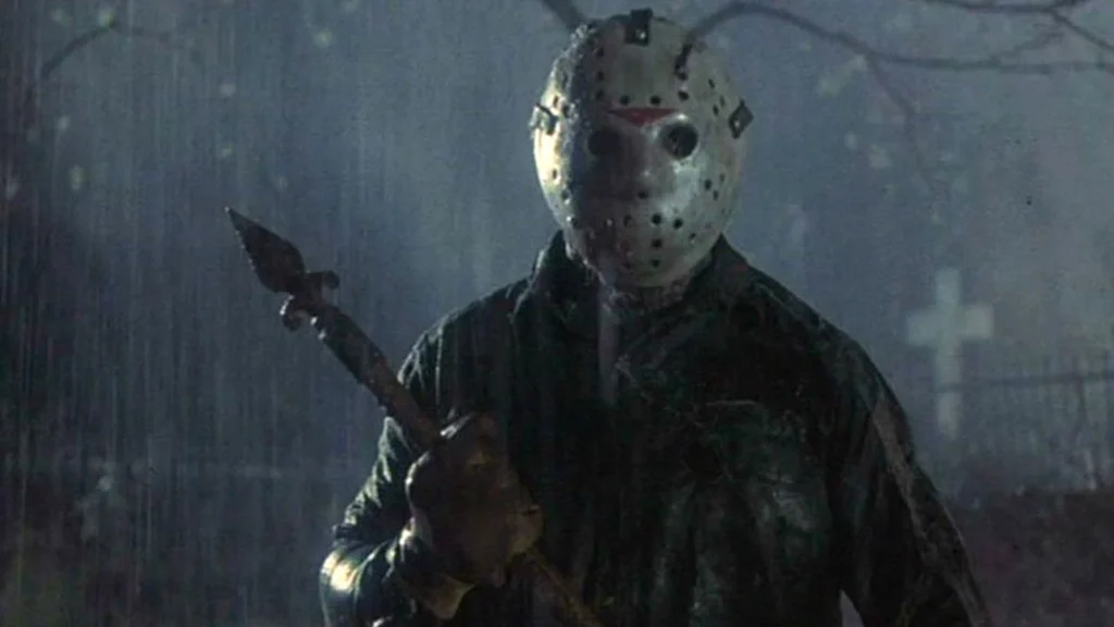 A menacing figure in a hockey mask stands in the rain at night, gripping a sharp spear-like weapon, with a graveyard cross faintly visible in the misty background. The eerie lighting and soaked setting heighten the horror atmosphere.