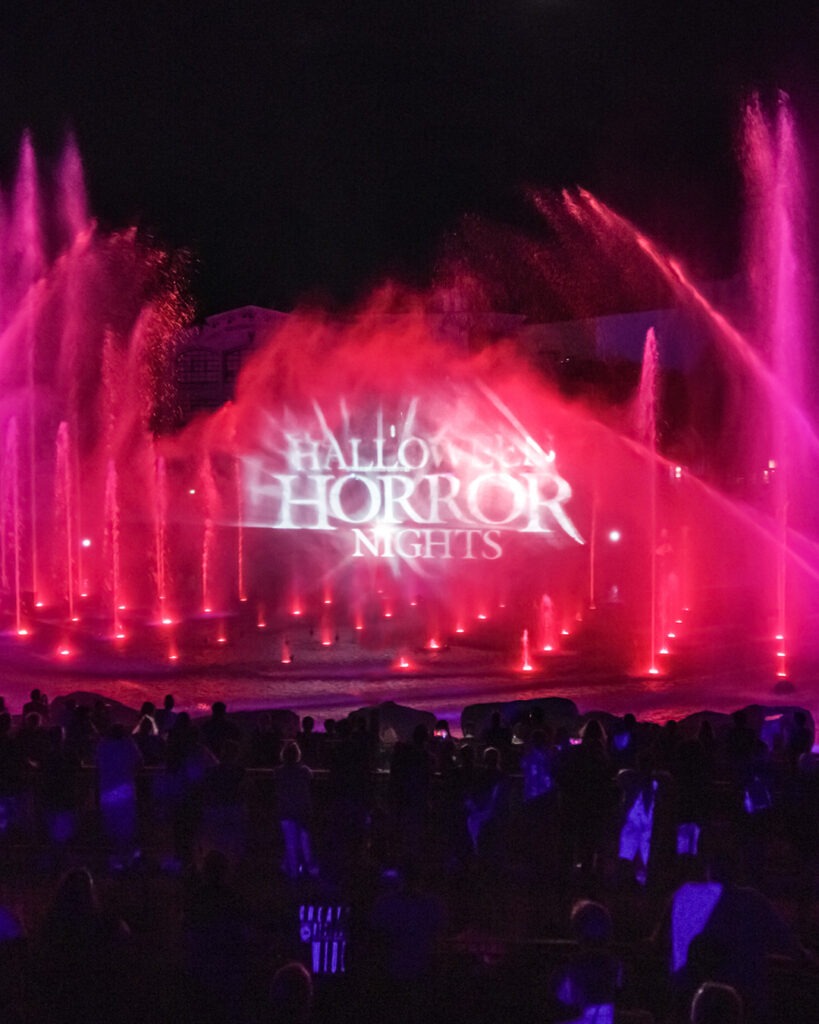 Nighttime water show at Halloween Horror Nights, featuring vibrant pink and red fountains framing a mist screen projection of the event's logo. Silhouetted spectators watch from the foreground, creating a dramatic and immersive Halloween atmosphere.