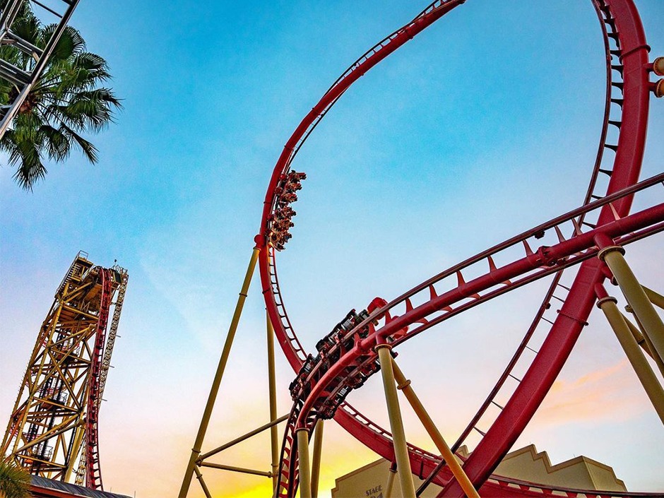 The red roller coaster track of Hollywood Rip, Ride, Rockit with riders speeding through a steep curve against a vibrant sunset sky. The towering vertical drop structure rises on the left, with palm trees framing the scene for a dramatic view.