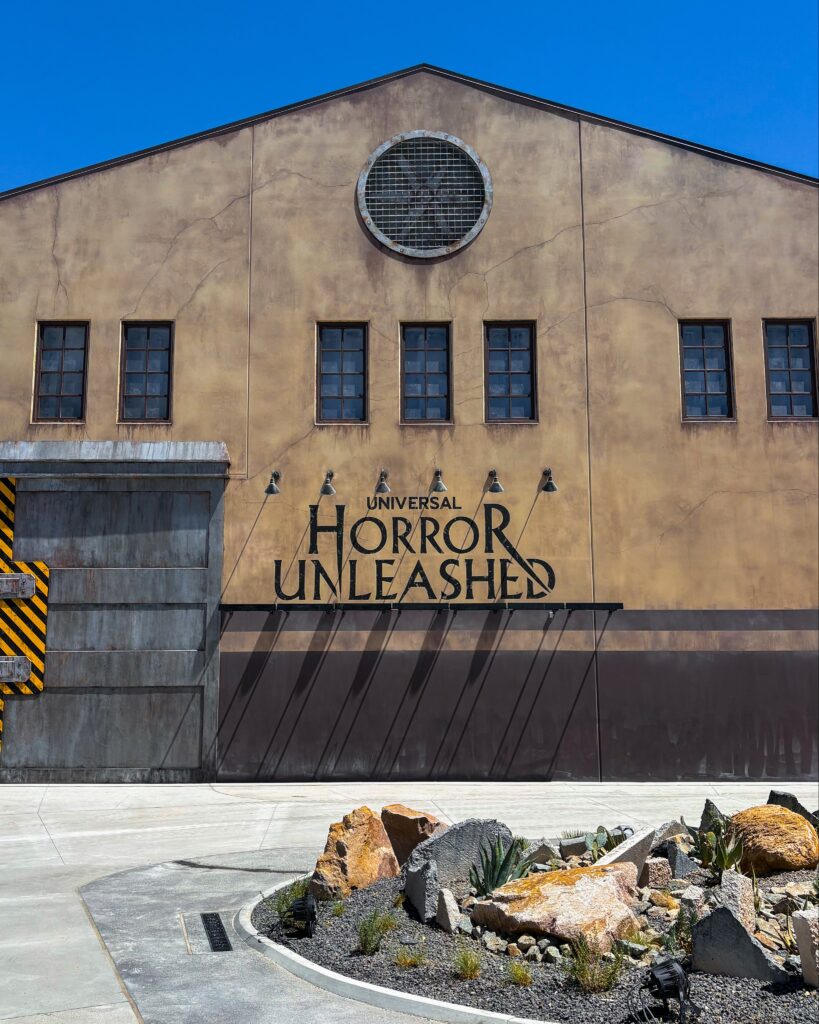The exterior of the “Universal Horror Unleashed” building is shown in daylight, with a weathered industrial design featuring cracked tan walls, black-framed windows, and a large circular vent near the roof peak. In front, a landscaped rock garden with desert plants contrasts against the stark concrete walkway.