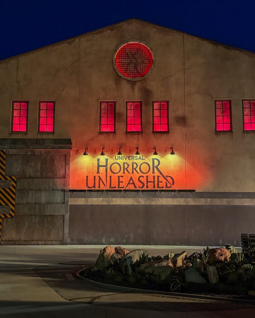 The “Universal Horror Unleashed” building glows at night with deep red light pouring from its windows and large circular vent, creating a foreboding atmosphere. The industrial-style facade is accented by weathered walls, warning-striped details, and a landscaped rock garden in the foreground.